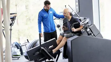 Eine Person trainiert an einem Trainingsgerät. Daneben steht ein Trainer.