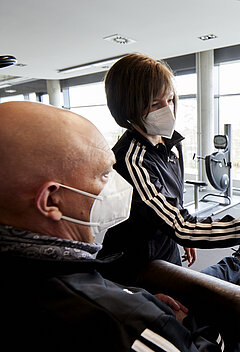 Eine Person trainiert an einem Trainingsgerät. Daneben steht eine Trainerin. Beide tragen einen Mund-Nasen Schutz.