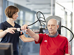 Eine Person trainiert an einem Trainingsgerät. Daneben steht eine Trainerin und leitet sie an.